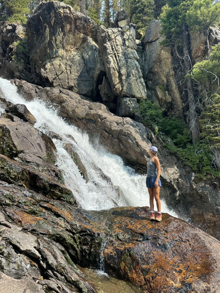 Kendyll standing on rocks beside a rushing waterfall in the mountains